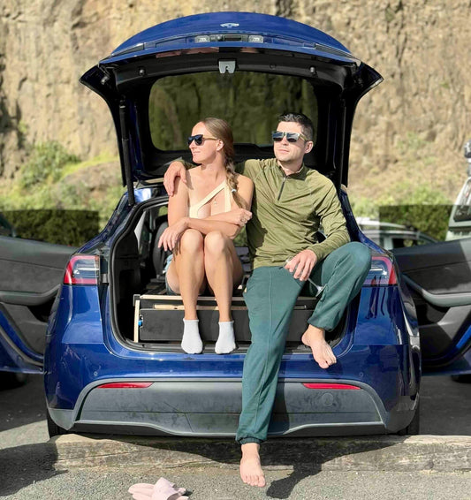 Couple sitting on a Teraglide platform bed inside a Tesla trunk, relaxing after a hike with scenic mountain backdrop