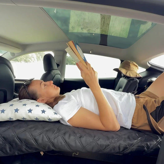 Person relaxing inside a Tesla Model Y on a Teraglide camping platform, reading a book and showing the headroom above the bed under the glass roof
