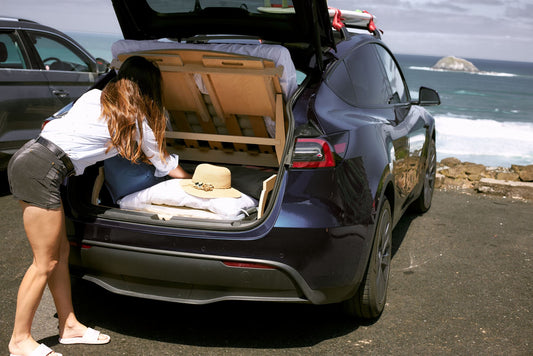 A woman organizing bedding under a lifted wooden camping platform in the trunk of a Tesla Model Y, parked by the ocean with waves in the background