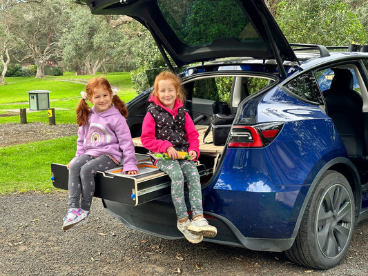 Two children sitting on a Teraglide platform bed in the trunk of a Tesla, enjoying family camping in a scenic outdoor setting