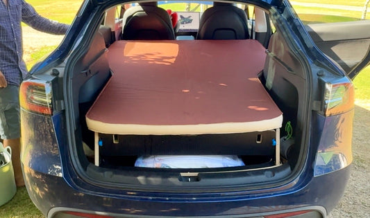 Tesla Model Y with Teraglide PRO camping platform and mattress set up in the open trunk at a campsite, showing a flat bed and storage space underneath