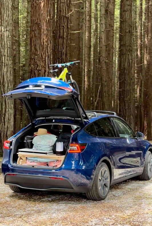 Tesla with open trunk, fully equipped Teraglide camping platform and gear, parked in a forest setting for an outdoor adventure