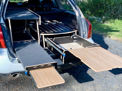 A platform bed designed to fit inside the trunk of a car, with wood surfaces and a plywood frame, intended for use in vehicle camping.