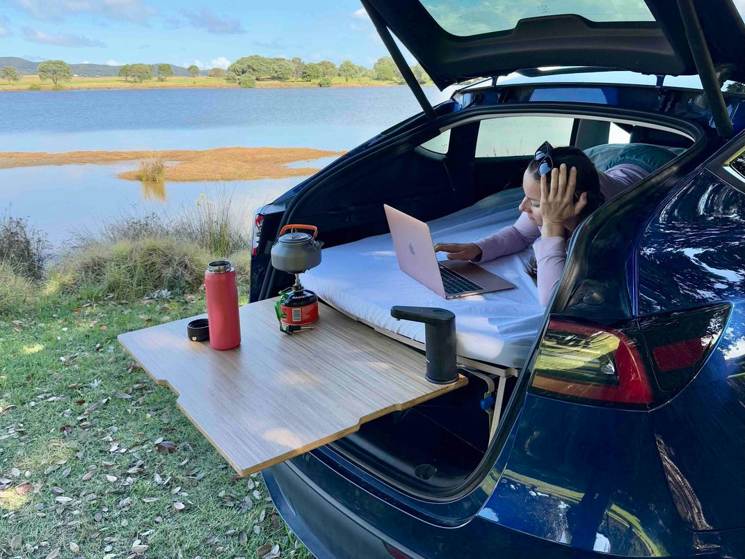 Woman working on a laptop on a Teraglide sleeping platform in a Tesla Model Y, with clip-on bamboo table and stove by a lakeside.